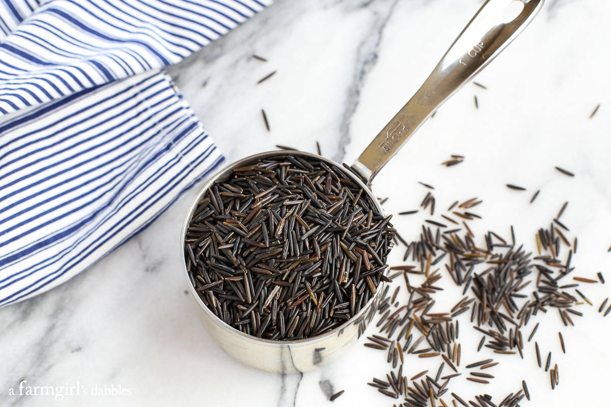 A measuring cup filled with wild rice, surrounded by scattered rice grains on a countertop.