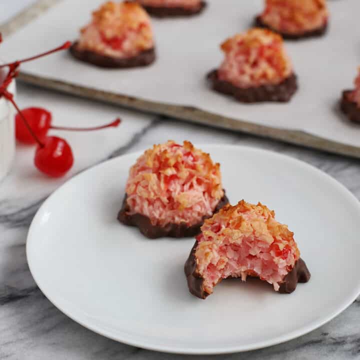 Two chocolate dipped cherry macaroons on a white plate, one with a bite missing