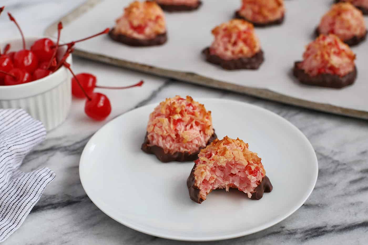 Two chocolate dipped cherry macaroons on a white plate, one with a bite missing