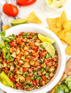 A white bowl of grilled shrimp ceviche garnished with lime wedges and tortilla chips