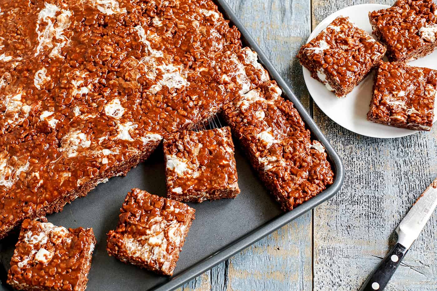 A tray of marshmallow chocolate peanut butter rice krispies bars, cut into small pieces with several on a plate.