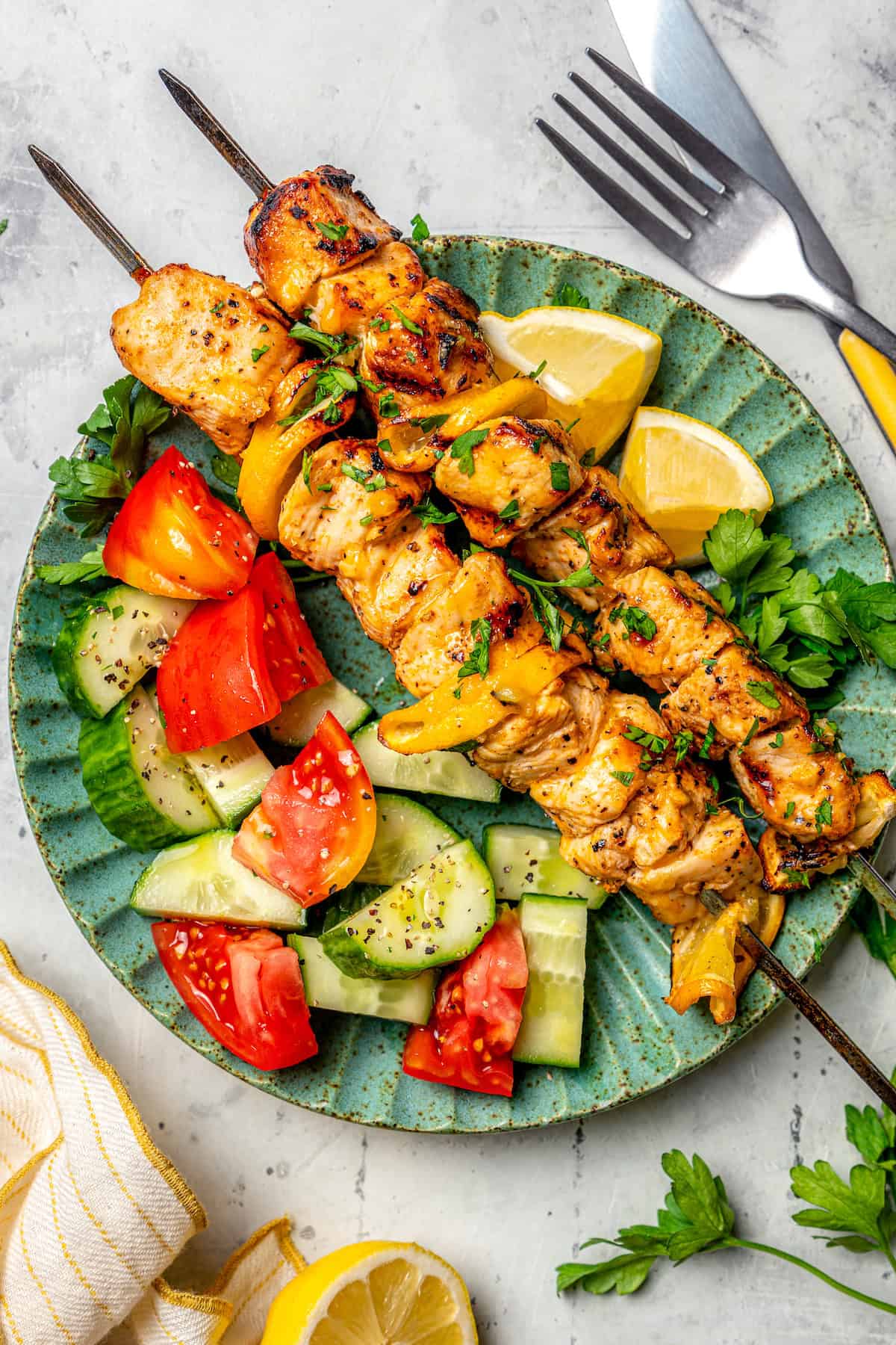 These Grilled Lemon Pepper Chicken Skewers are an easy summer recipe that's super fast for a weeknight. Don't skip the lemon butter sauce! Overhead view of grilled lemon pepper chicken skewers on plate with cucumbers, tomatoes, and lemon wedges