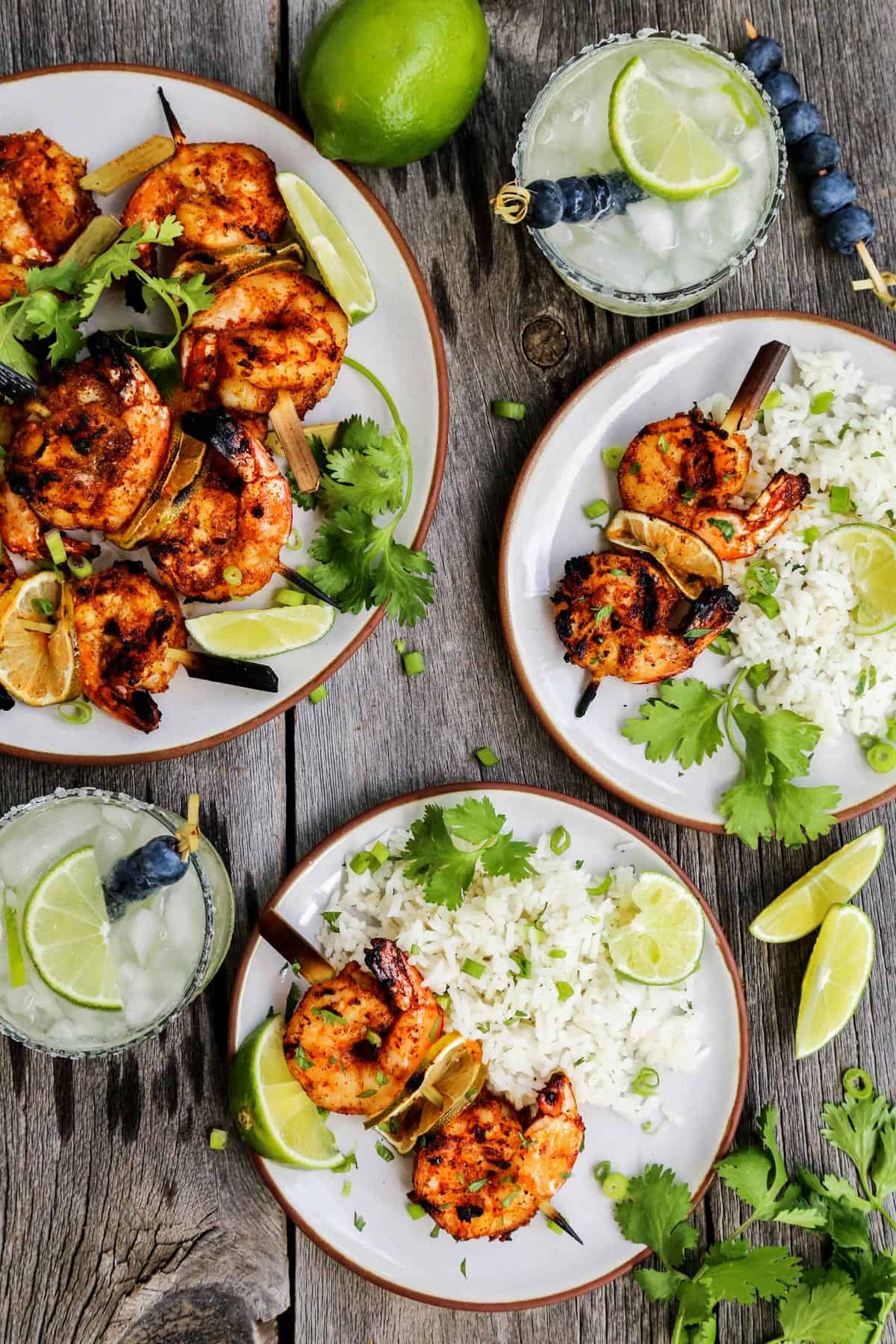 Grilled shrimp skewers on plates with white rice and fresh limes, also glasses of margaritas.