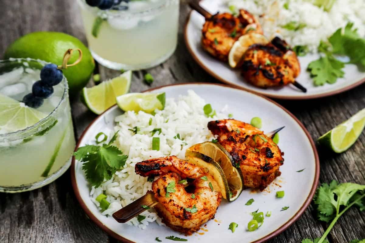 small plates of grilled shrimp skewers with white rice and fresh limes, also glasses of margaritas