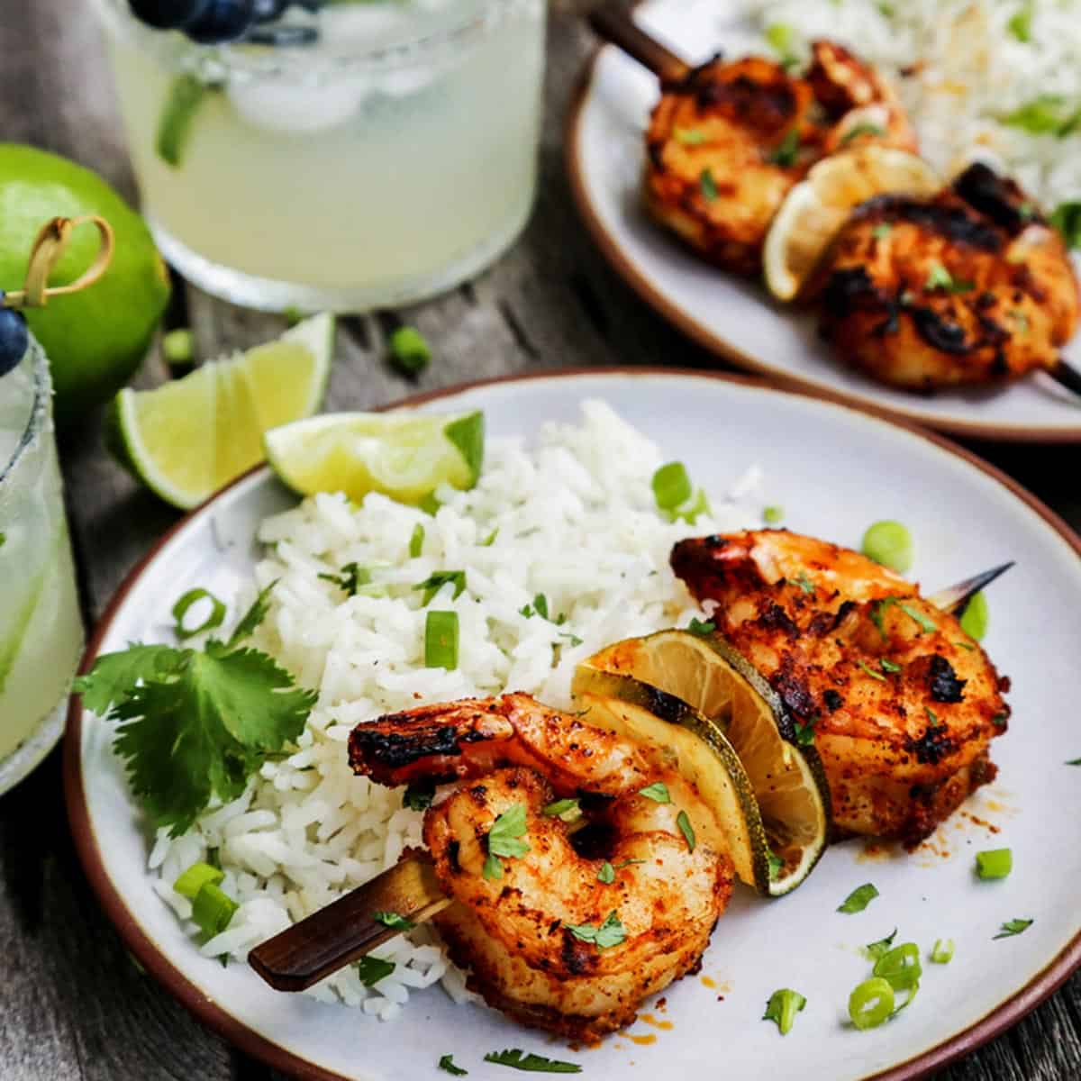 grilled shrimp skewers on plates with white rice and fresh limes, also glasses of margaritas