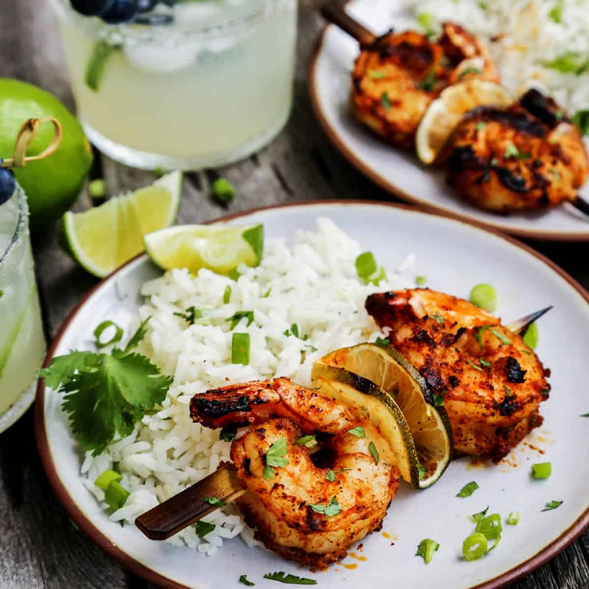 Flavorful margarita grilled shrimp kabobs are charred to perfection. This grilled shrimp recipe is perfect for summer dinners and appetizers! Grilled shrimp skewers on plates with white rice and fresh limes, also glasses of margaritas.