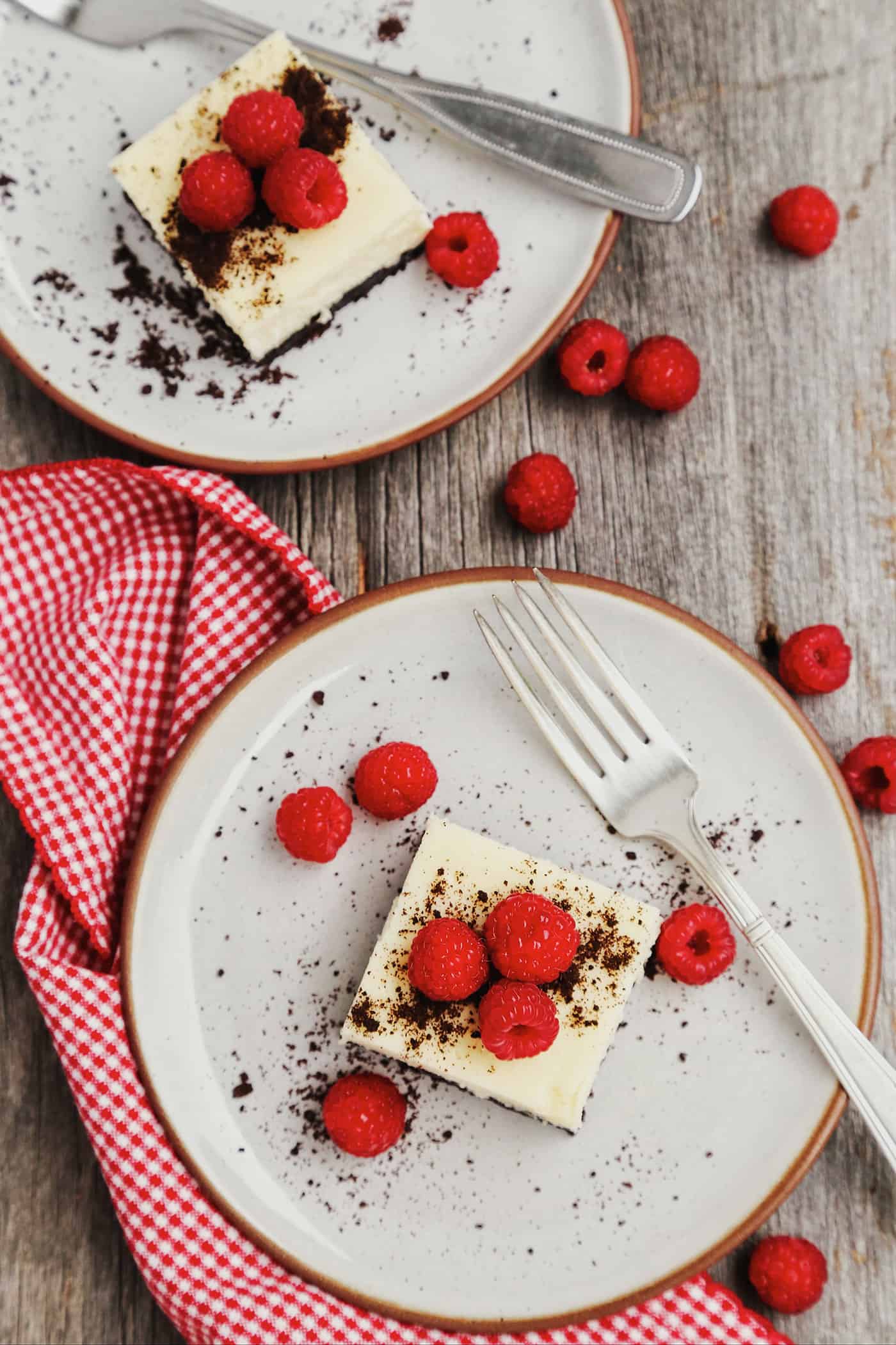 Overhead view of two cheesecake squares on white plates topped with raspberries