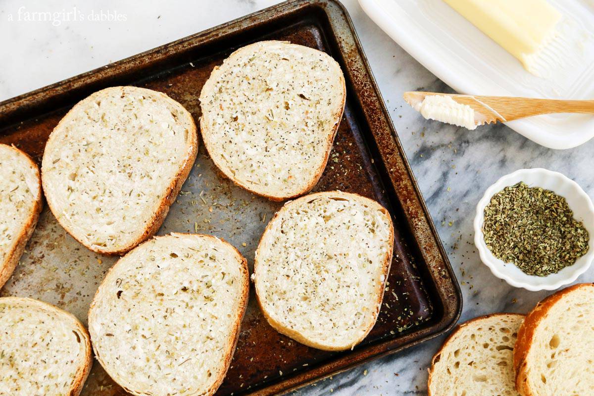 Landscape photo of sliced bread on a baking sheet, with butter on the side for spreading and a small bowl of Italian seasoning.