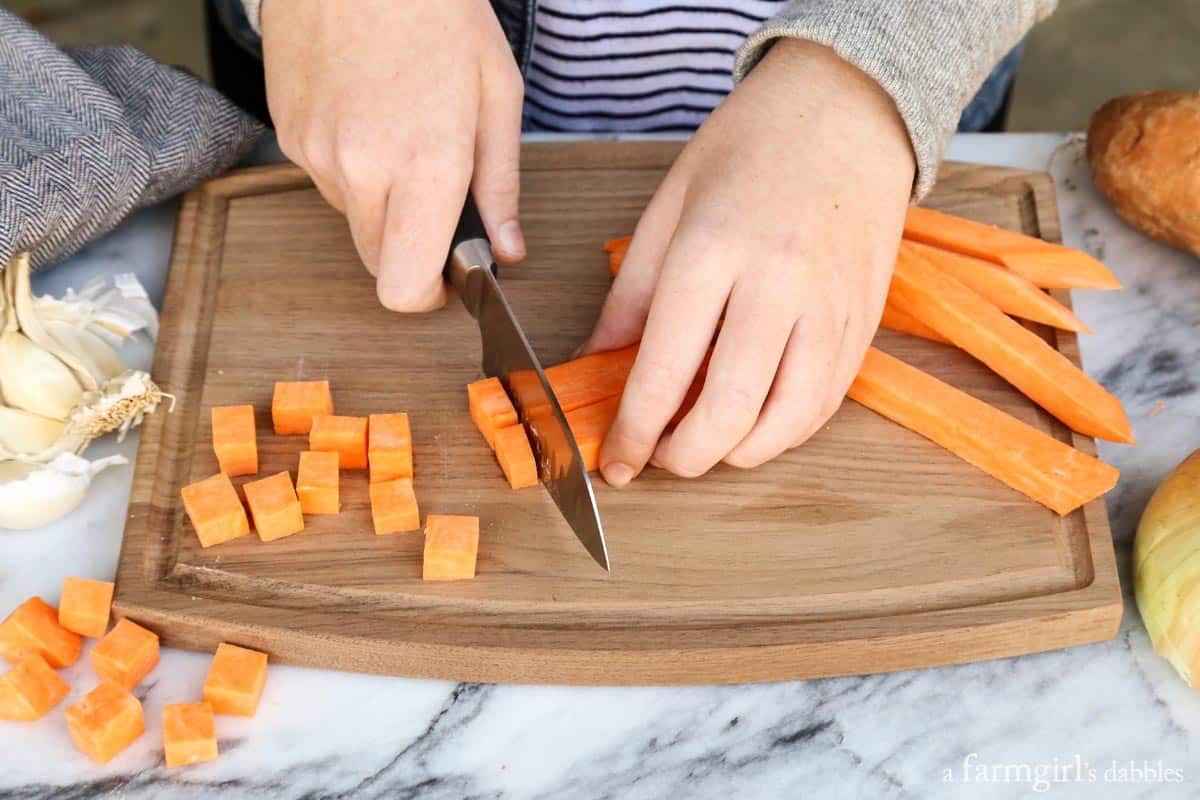 Hands using a large knife to cut sweet potatoes into cubes on a wooden cutting board.
