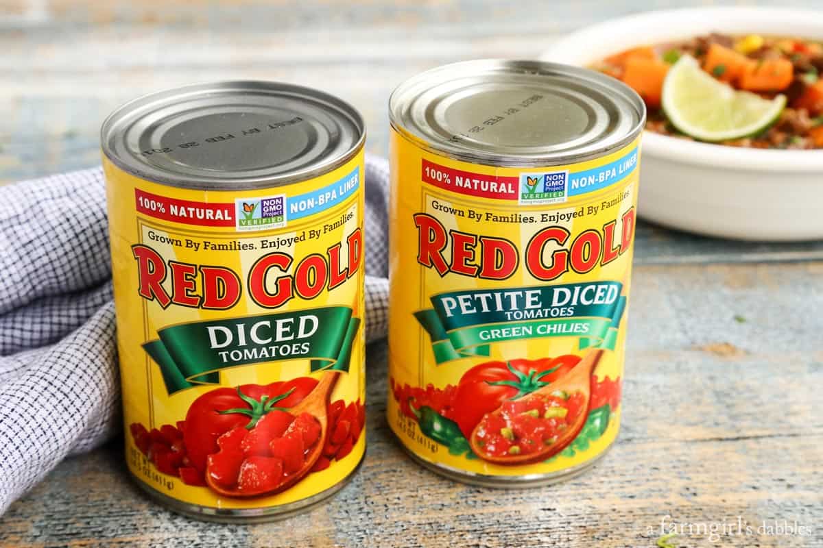 Two cans of Red Gold tomatoes on a countertop with a bowl of stew in the background.