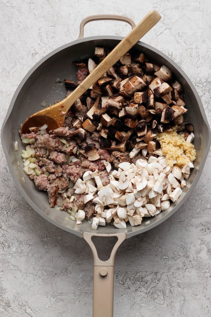 Wooden spoon stirring mushrooms and garlic into pan of browned sausage.