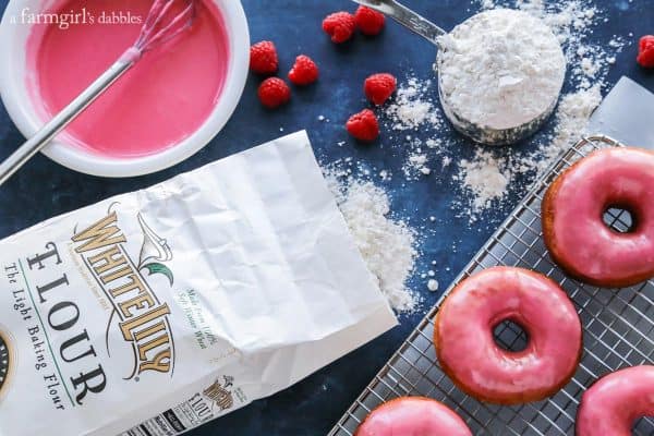 Yeast Donuts with Raspberry Glaze surrounded by flour, raspberries and a bowl of glaze