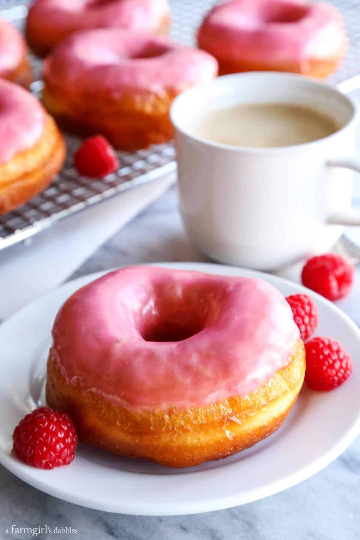 This golden, vanilla-raspberry donuts recipe with sweet lemon glaze is a yummy homemade gift for Valentine's Day. Homemade raspberry donuts with a cup of coffee in the background.