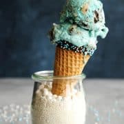 Cookie Monster Ice Cream in a dipped cone propped up inside a glass filled with white sprinkles.