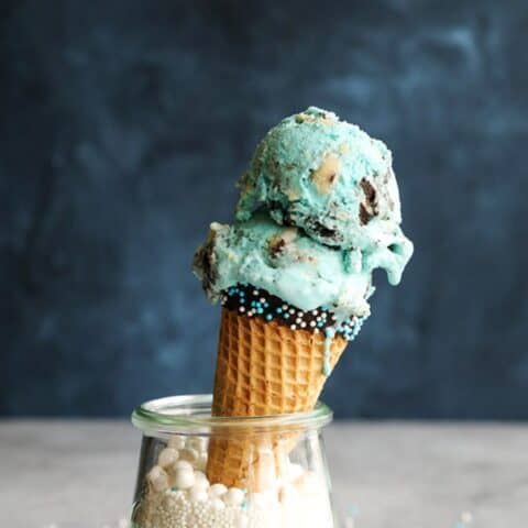 Cookie Monster Ice Cream in a dipped cone propped up inside a glass filled with white sprinkles.