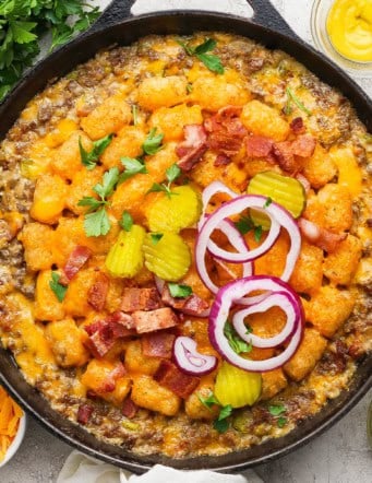 Bacon cheeseburger tater tot casserole in skillet with pickles and onions on top.
