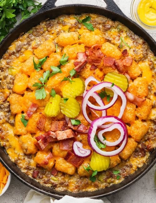 Bacon cheeseburger tater tot casserole in skillet with pickles and onions on top.