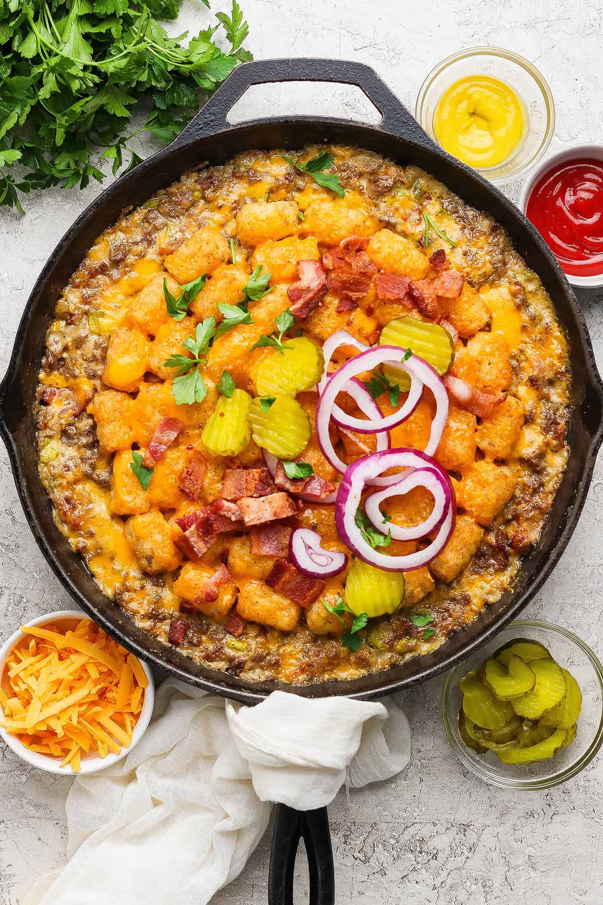 Bacon cheeseburger tater tot casserole in skillet with pickles and onions on top.