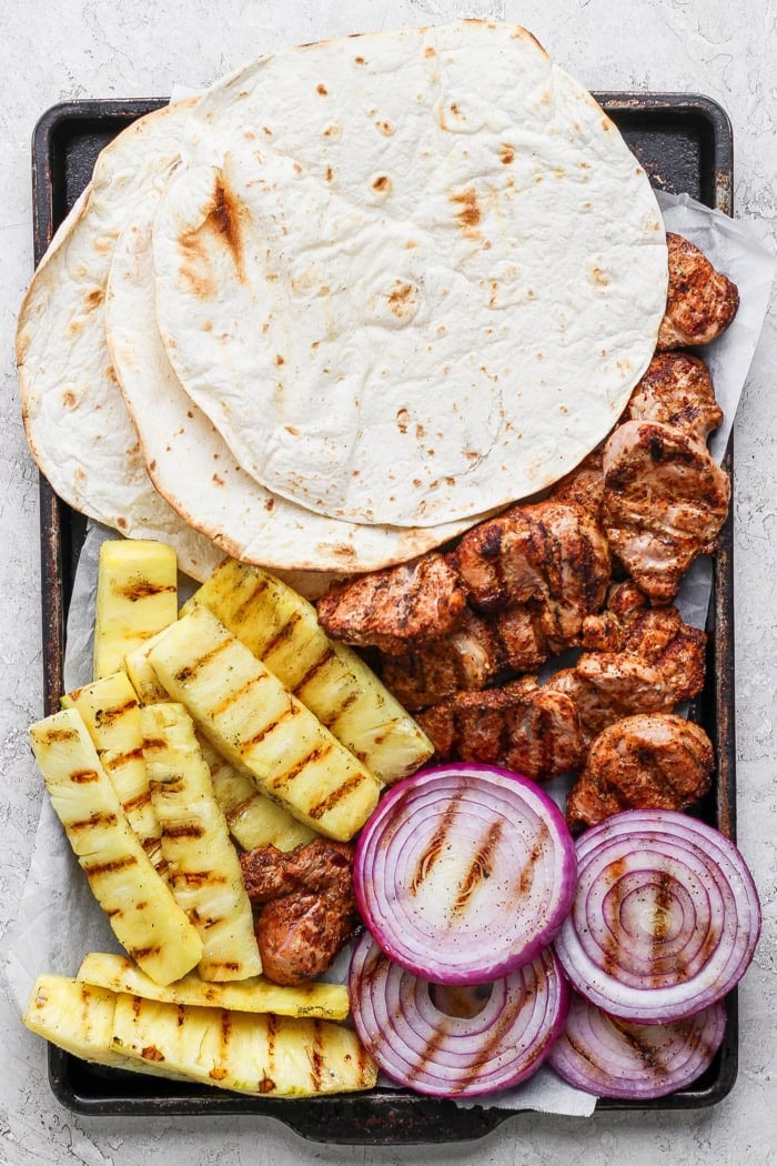 Overhead view of flour tortillas, grilled pork, pineapple, and red onion slices on sheet pan
