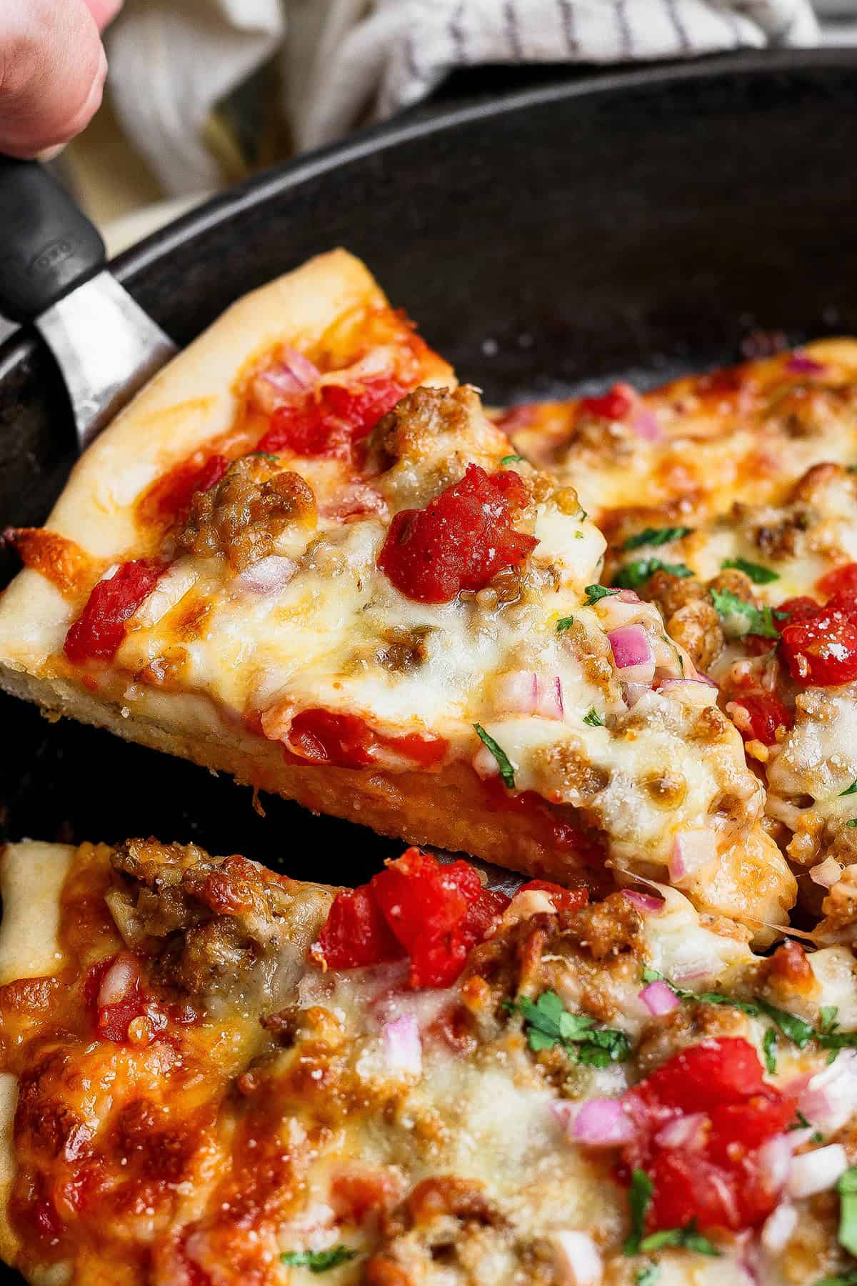 This skillet pizza with sausage has an Asian-inspired twist with chili garlic tomato sauce! A cast iron favorite with a crisp, chewy crust. Lifting slice of skillet pizza from pan.