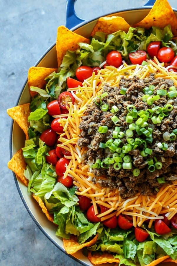 Dorito taco salad layered in a large blue bowl, with nacho cheese Doritos, lettuce, tomatoes, cheese, and taco meat