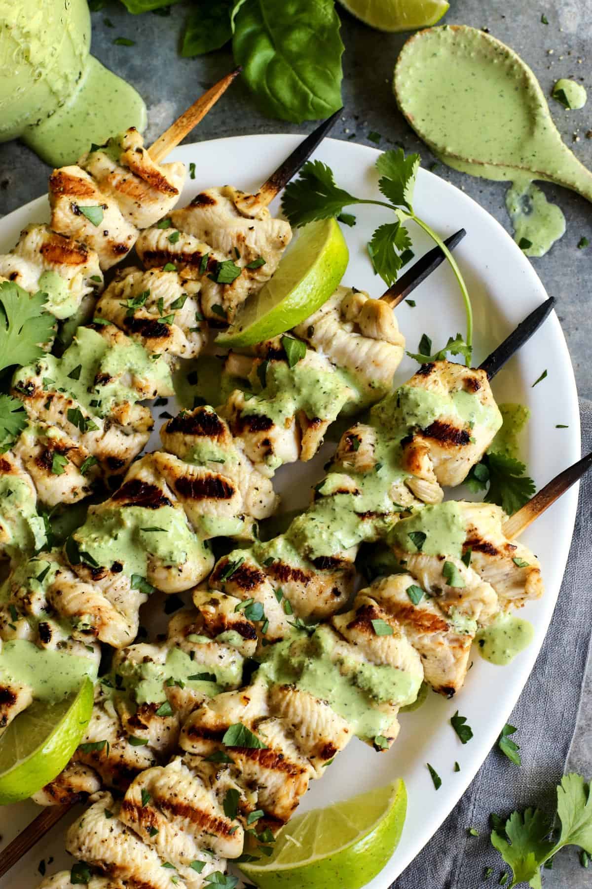 These grilled chicken skewers pair tender marinated strips of grilled chicken with a tangy green curry sauce. The perfect summer dinner! Overhead view of grilled chicken skewers on platter with green curry yogurt sauce