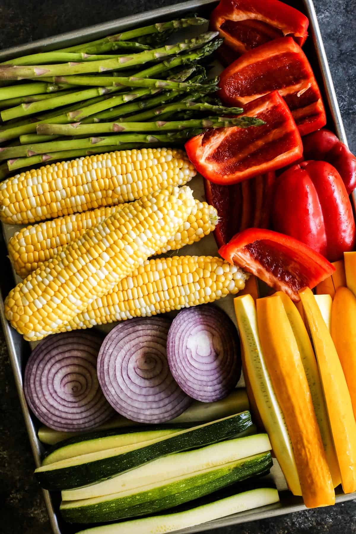 fresh vegetables on a rimmed pan: asparagus, sweet corn, and sliced red pepper, yellow squash, zucchini, and red onion
