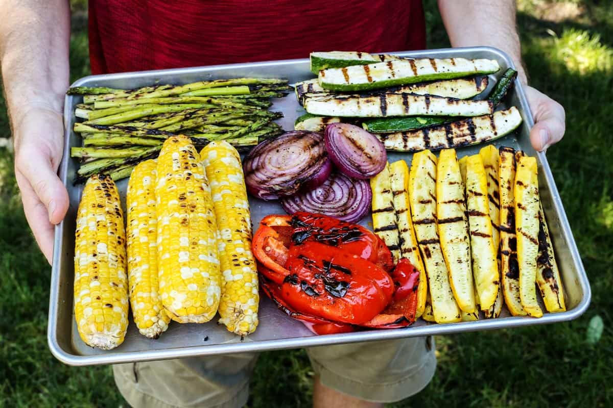 man holding rimmed pan of grilled vegetables: asparagus, sweet corn, red pepper, red onion, yellow squash, zucchini