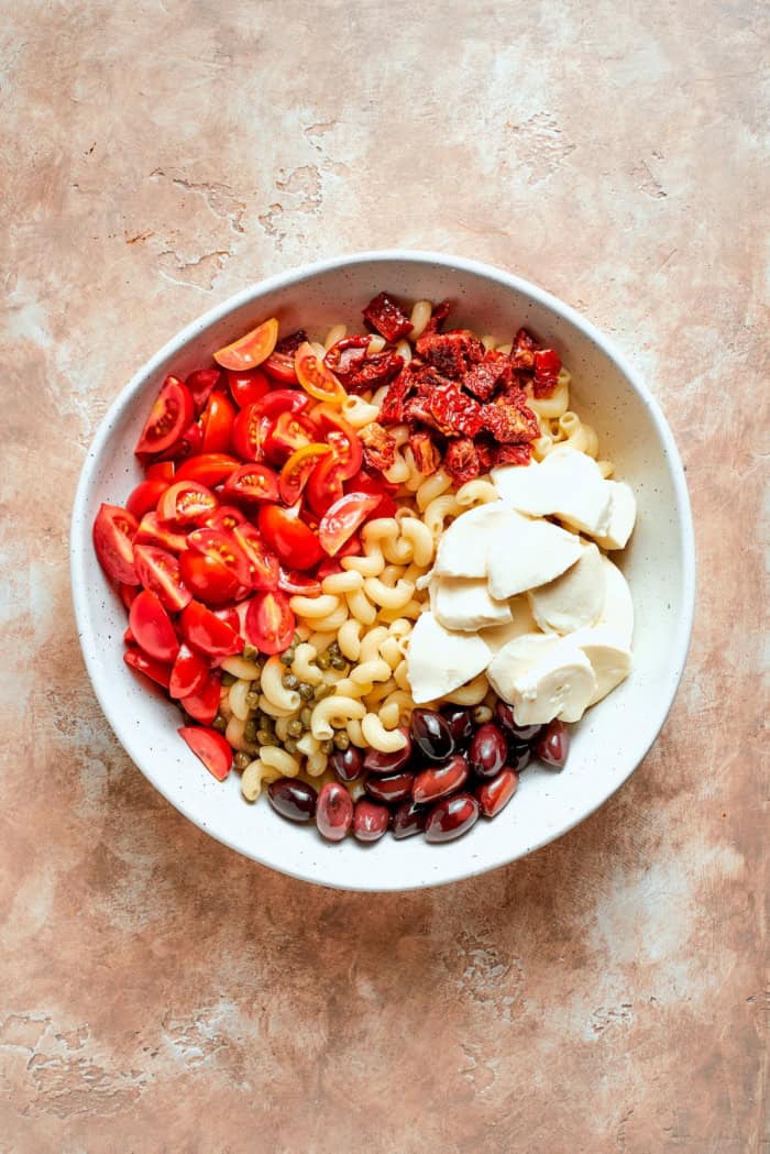 This sun-dried tomato pasta salad lets the abundance of summer produce shine, with juicy tomatoes and fresh basil. Perfect for picnics! Pasta, olives, mozzarella, tomatoes, and sun-dried tomatoes in bowl.