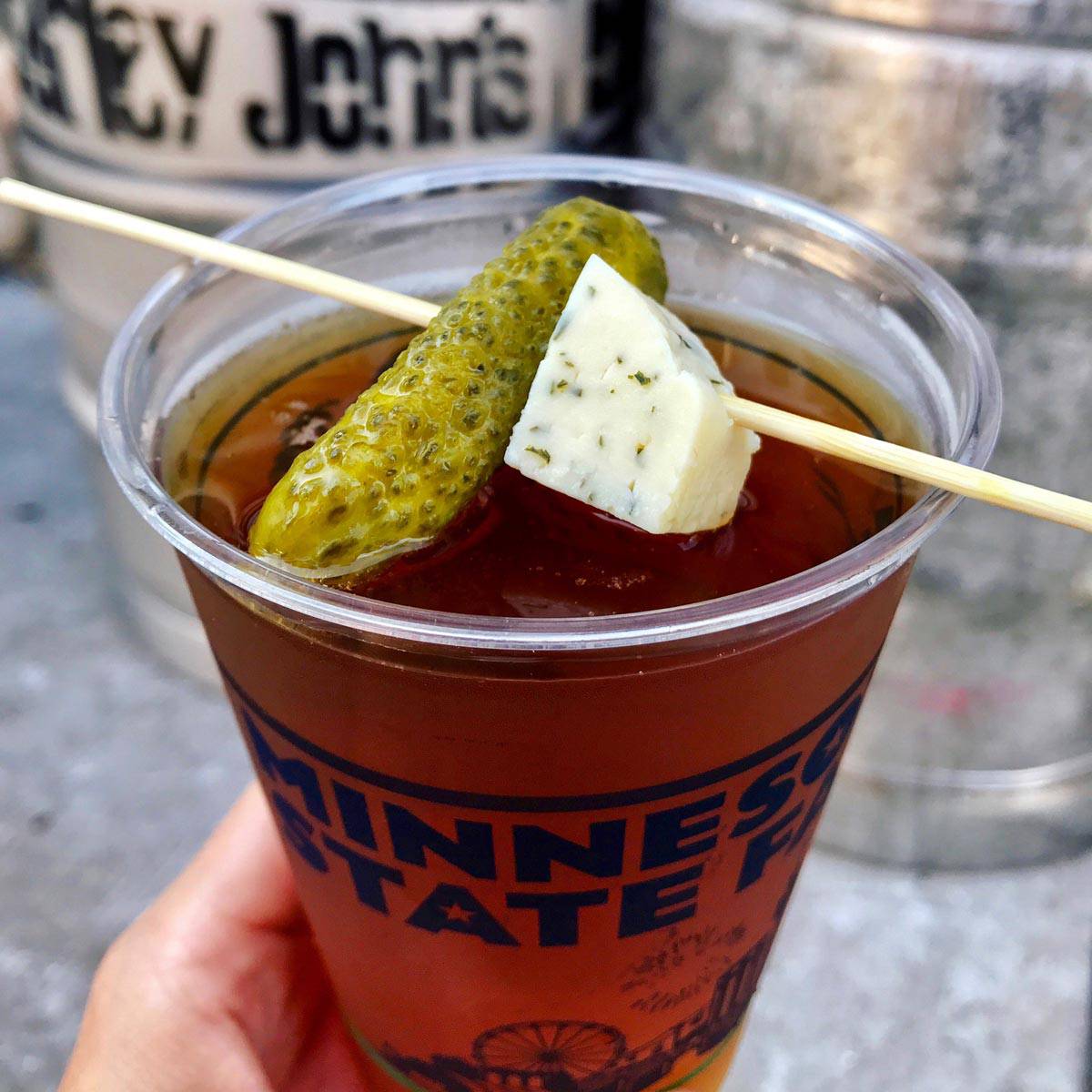 a glass of beer with a skewer on it with a pickle and cube of cheese