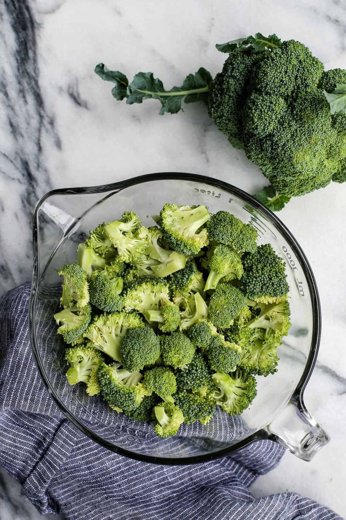 Overhead view of broccoli florets in a glass bowl next to a head of broccoli on a countertop.