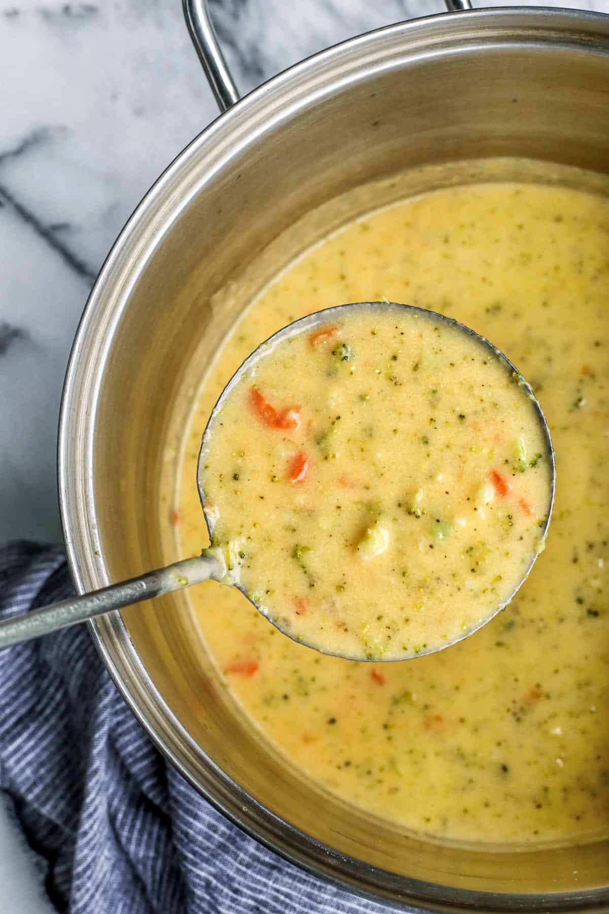 Overhead view of a ladle of broccoli cheddar soup held over the rest of the soup in a large pot.