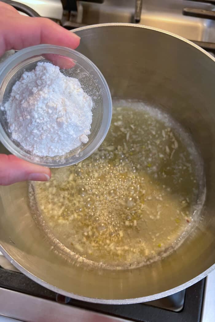 A hand holding a glass bowl of flour over a soup pan with butter