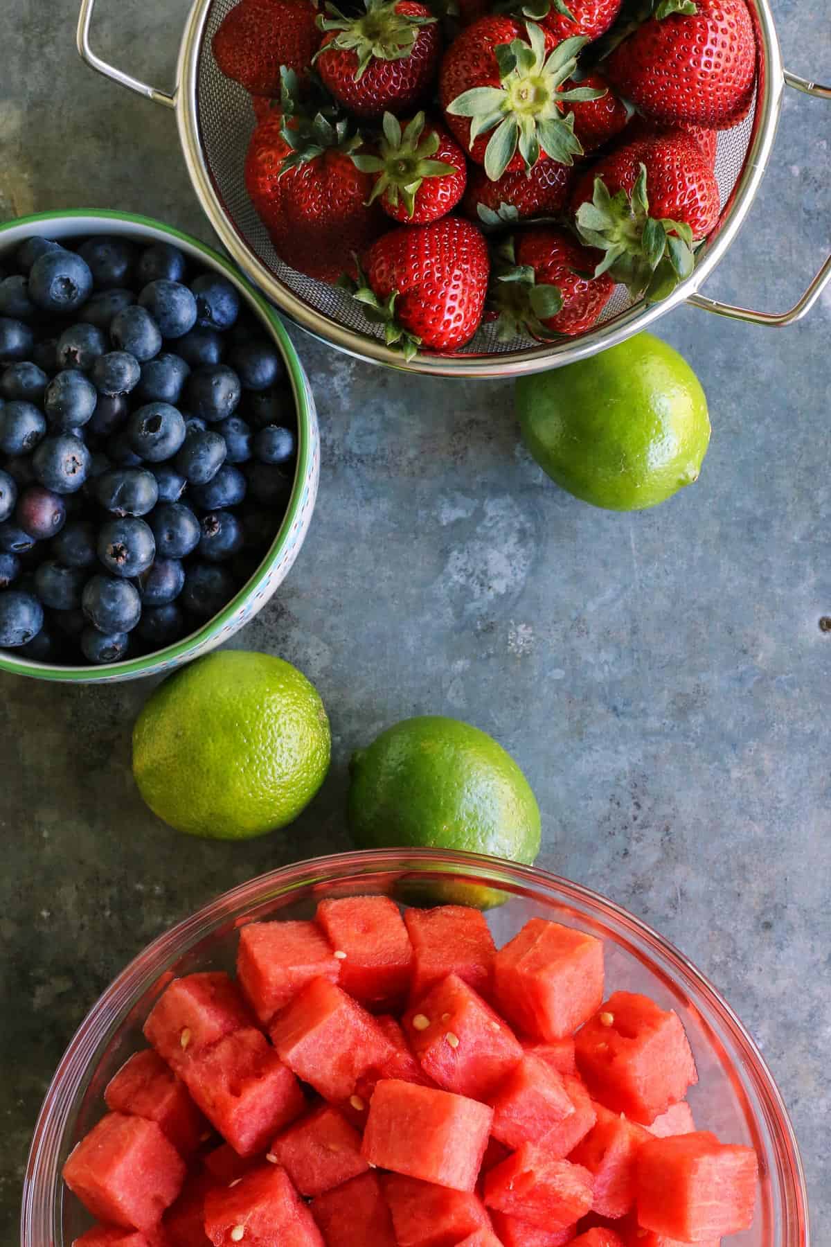 colander of strawberries, bowl of blueberries, bowl of watermelon, whole limes