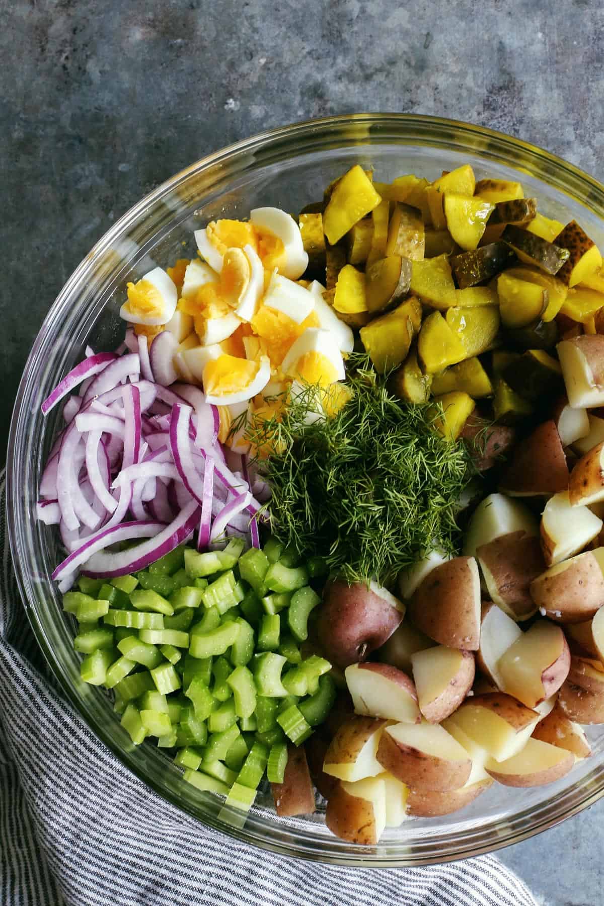 Potatoes, dill pickles, hard boiled eggs, red onions, celery, and dill in a clear bowl.