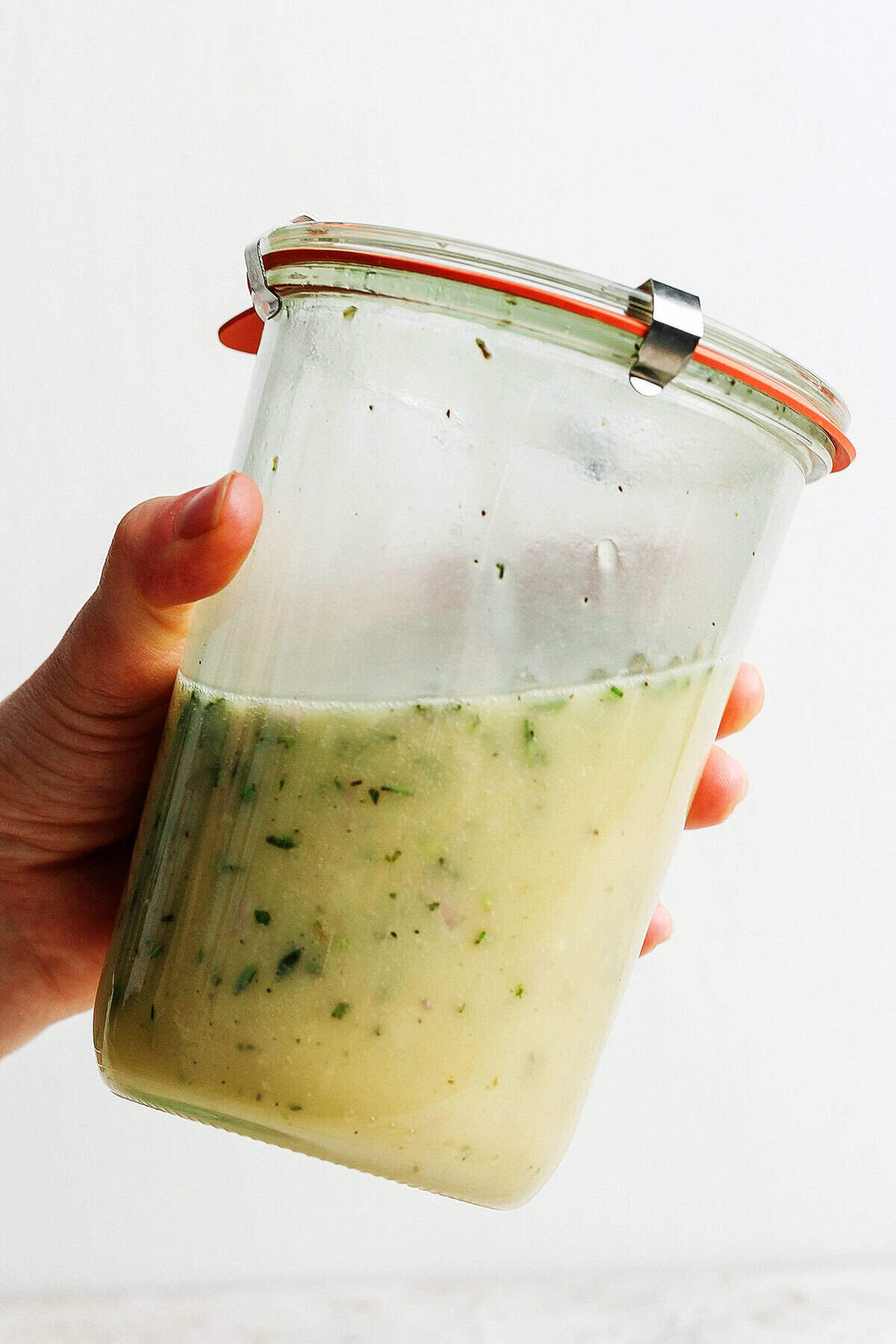 This homemade Lemon Vinaigrette recipe is loaded with bright flavor and fresh herbs. Use it on salads, grilled vegetables, and so much more! Hand holding jar with emulsified lemon vinaigrette
