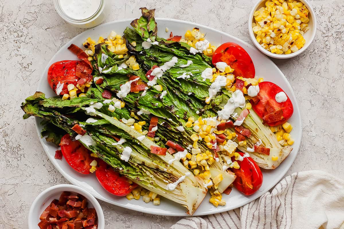 This smoky Grilled Romaine Salad is simple and summery, with tomatoes, sweet corn, crispy bacon and homemade buttermilk ranch dressing. Yum! Overhead view of grilled romaine salad on platter with grilled tomatoes and corn