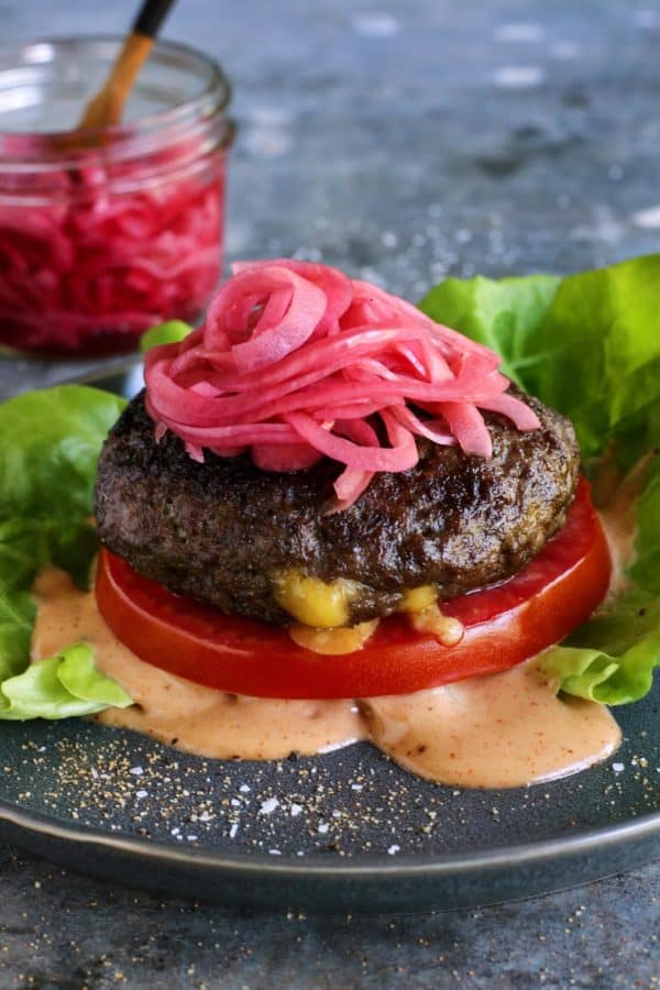 a Juicy Lucy cheese-stuffed burger on a plate, with lettuce, pickled red onions, slice of tomato, and creamy burger sauce