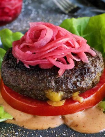a Juicy Lucy cheese-stuffed burger on a plate, with lettuce, pickled red onions, slice of tomato, and creamy burger sauce