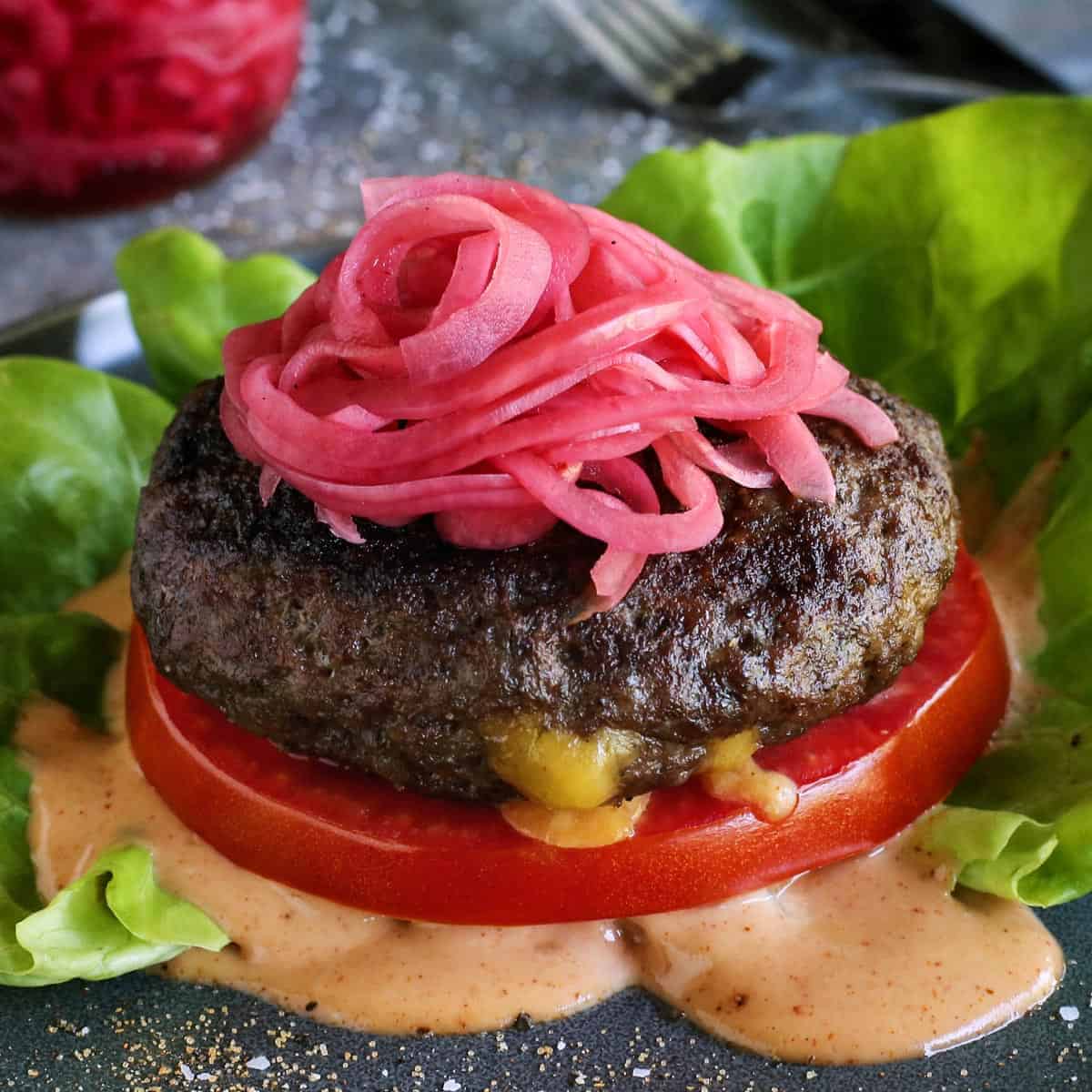 a Juicy Lucy cheese-stuffed burger on a plate, with lettuce, pickled red onions, slice of tomato, and creamy burger sauce