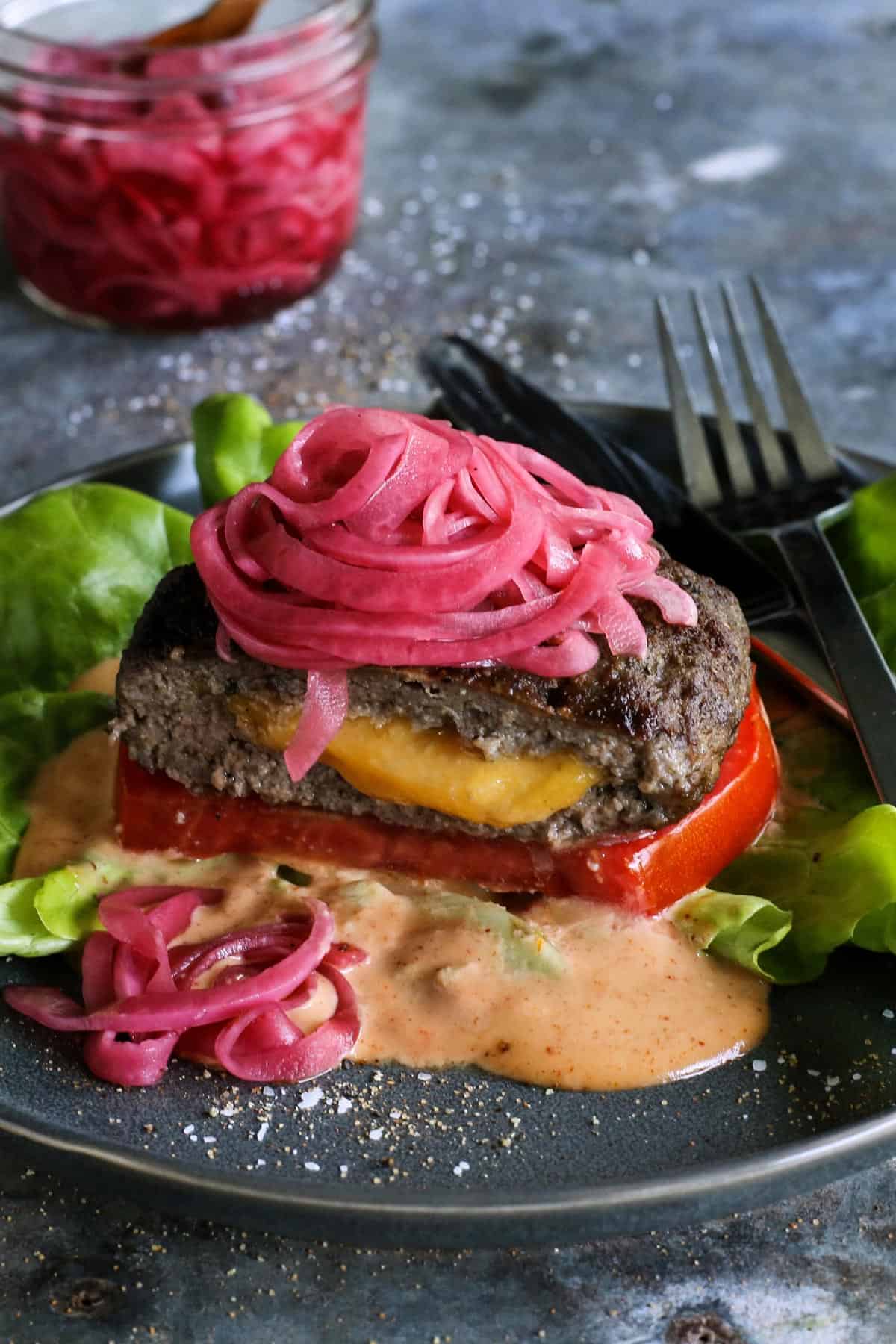 a Juicy Lucy cheese-stuffed burger on a plate, with lettuce, pickled red onions, slice of tomato, and creamy burger sauce