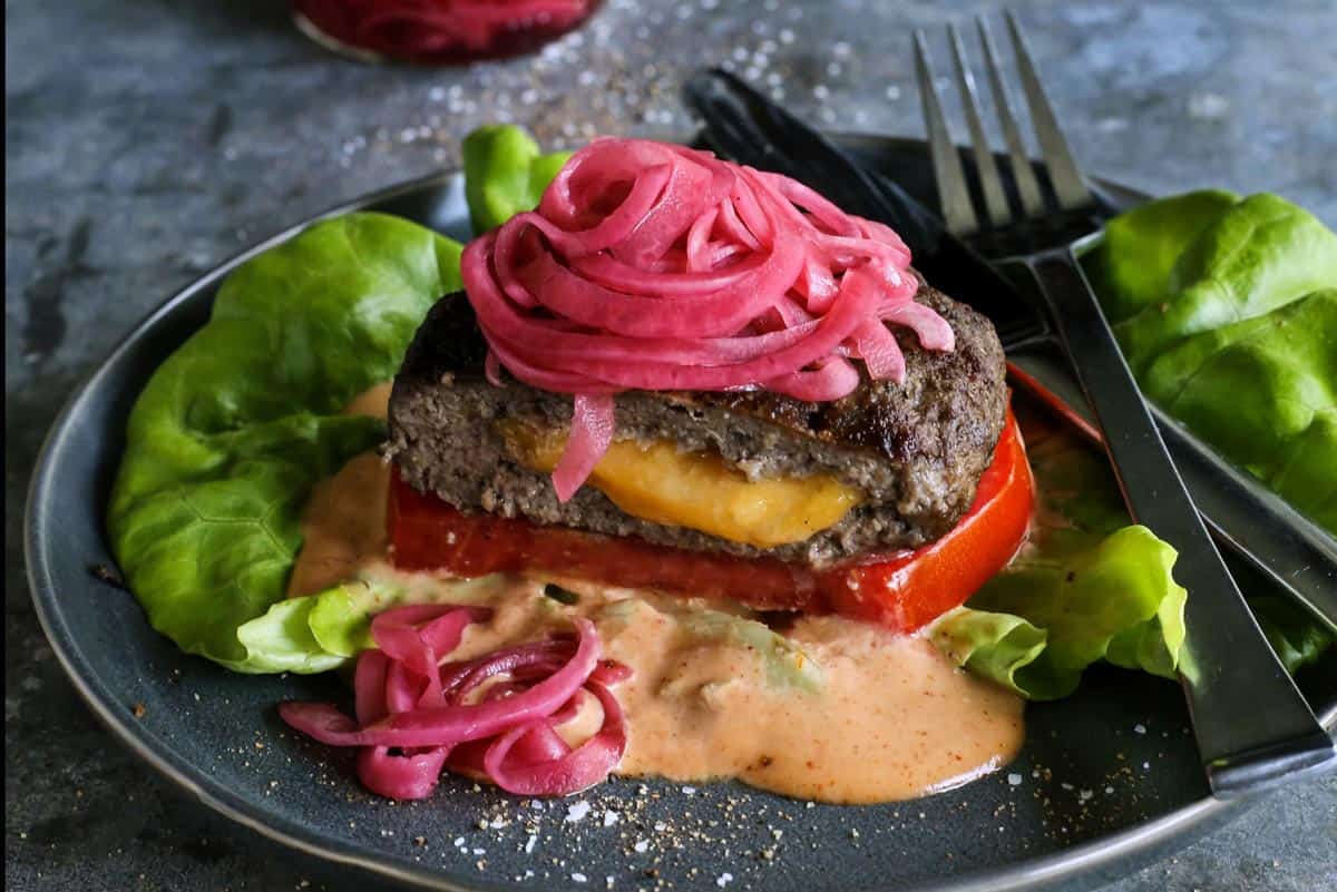 a Juicy Lucy cheese-stuffed burger on a plate, with lettuce, pickled red onions, slice of tomato, and creamy burger sauce