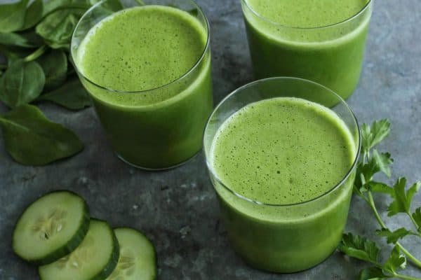 three glasses of green juice with spinach, cucumber slices, and parsley on the side