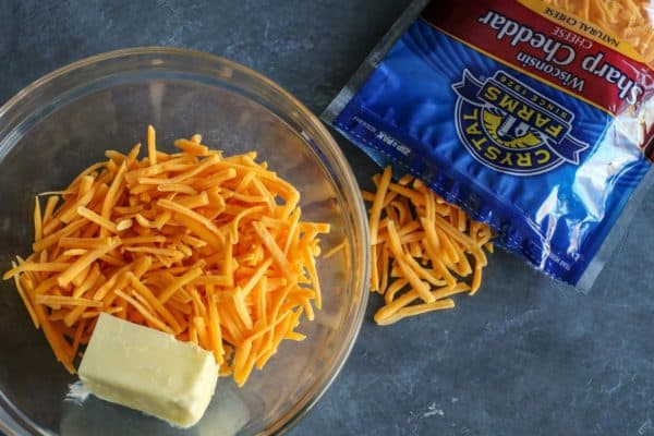 shredded sharp cheddar cheese and butter in a clear bowl, a package of Crystal Farms Sharp Cheddar on the side