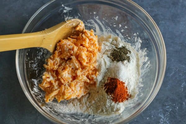 shredded sharp cheddar cheese, butter, flour, paprika, salt, and black pepper in a clear bowl with a wooden spoon