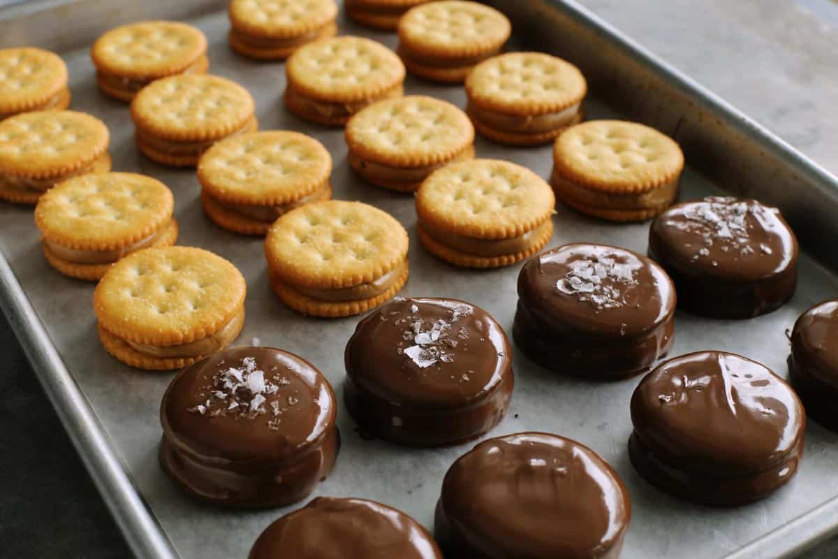 Ritz sandwich cookies with peanut butter inside, some are dipped in chocolate, some are sprinkled with sea salt flakes, all lined up on a rimmed pan