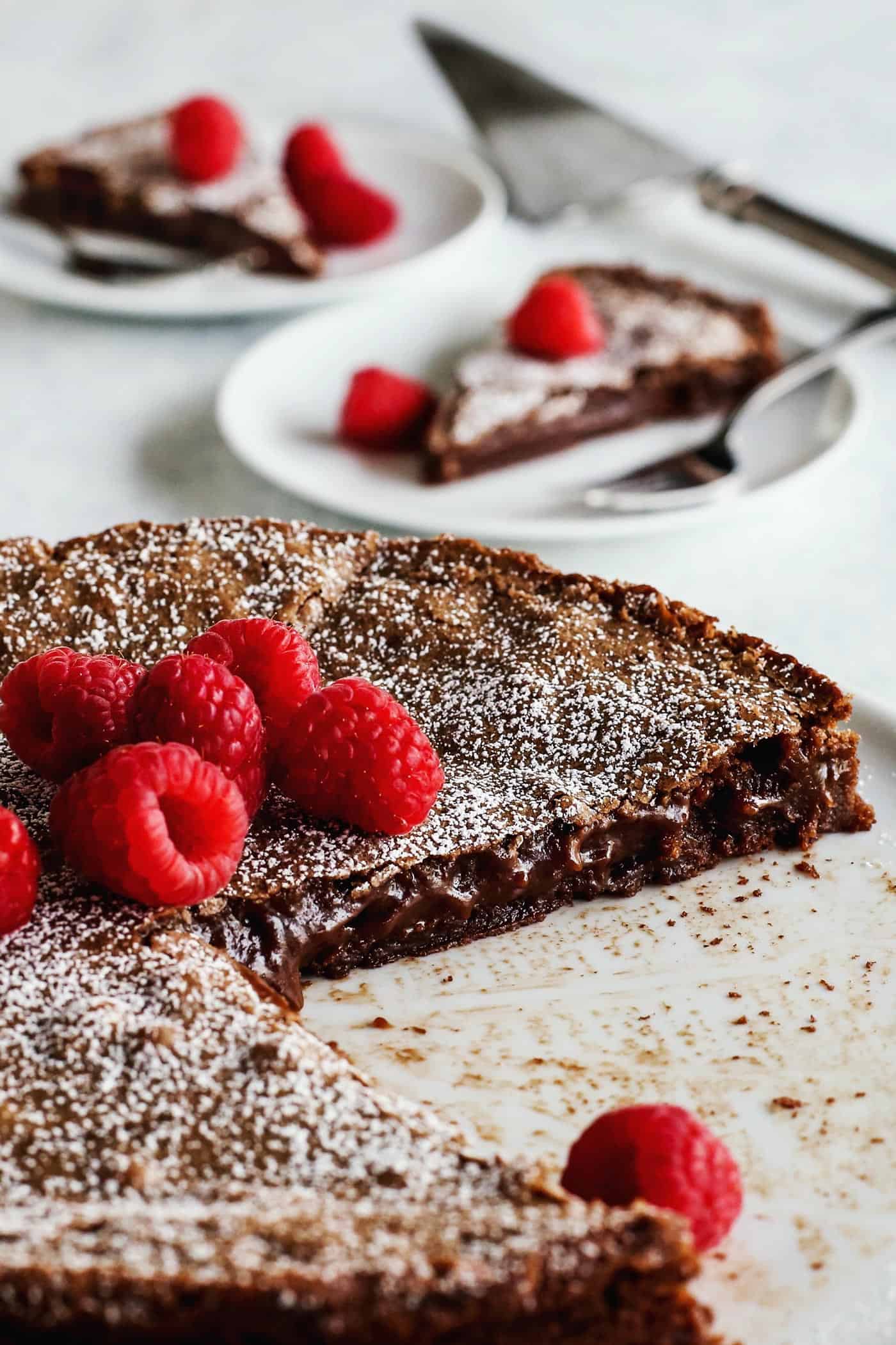 Swedish Kladdkaka chocolate cake on white cake plate, topped with fresh raspberries. Two pieces of cake on individual plates in the background.