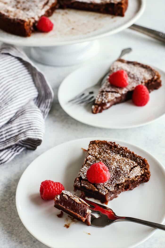 two individual plates with slices of chocolate cake and fresh raspberries, plus a cake platter with the cake on it