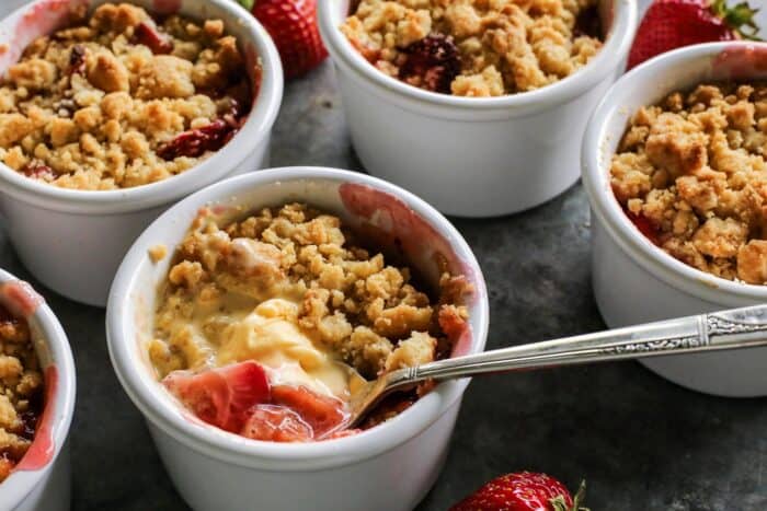 a spoon dipping into melty vanilla ice cream in a ramekin of rhubarb and strawberry crumble