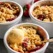 four white ramekins of strawberry rhubarb crumble, one with a scoop of ice cream and a spoon